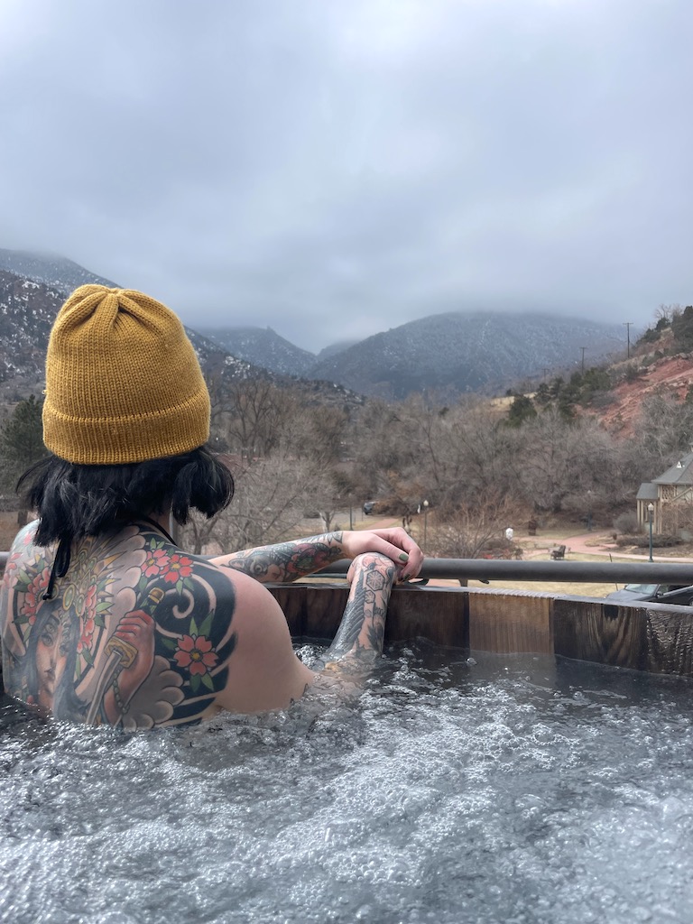 Woman with colorful tattoos and yellow beanie hat sitting in outdoor wooden hot tub with bubbling mineral water overlooking mountains and park on cloudy, snowy winter day. SunWater Spa, Manitou Springs, Colorado Springs, Colorado.