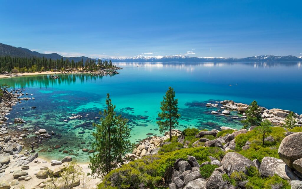 Rocky shoreline surrounded by pine trees overlooking bright blue/green clear water of Lake Tahoe with snow-capped mountains in distance on sunny day. Lake Tahoe, California, Nevada.