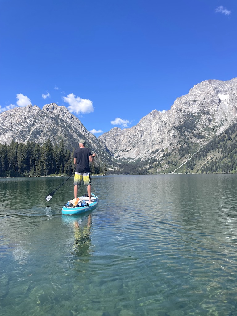 Man stand up paddle boarding on clear mountain lake with dramatic mountain views in immediate background on sunny summer day. Stand up paddle boarding Leigh Lake, Grand Teton National Park, Wyoming.