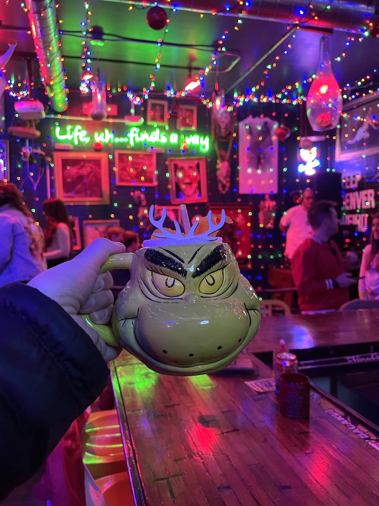 Person holding green Grinch mug with colorful, festive holiday lights and bar top in background at night. Electric Cure Krampus Room holiday pop up, Denver, Sloan's Lake, Colorado.