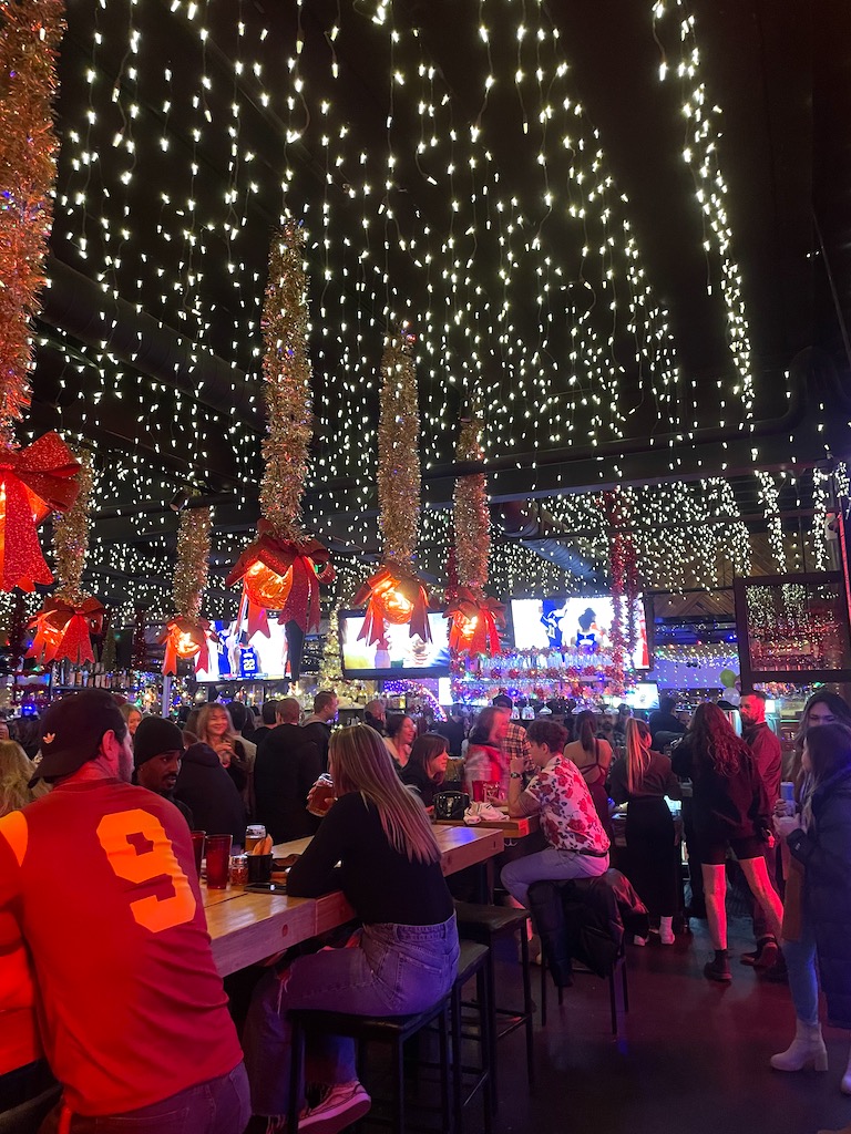Huge string lights and bows hanging from tall ceiling above large bar full of people at night. Happy Camper Pizza decorated for Christmas, Happy Camper, Denver, Highlands, Colorado.