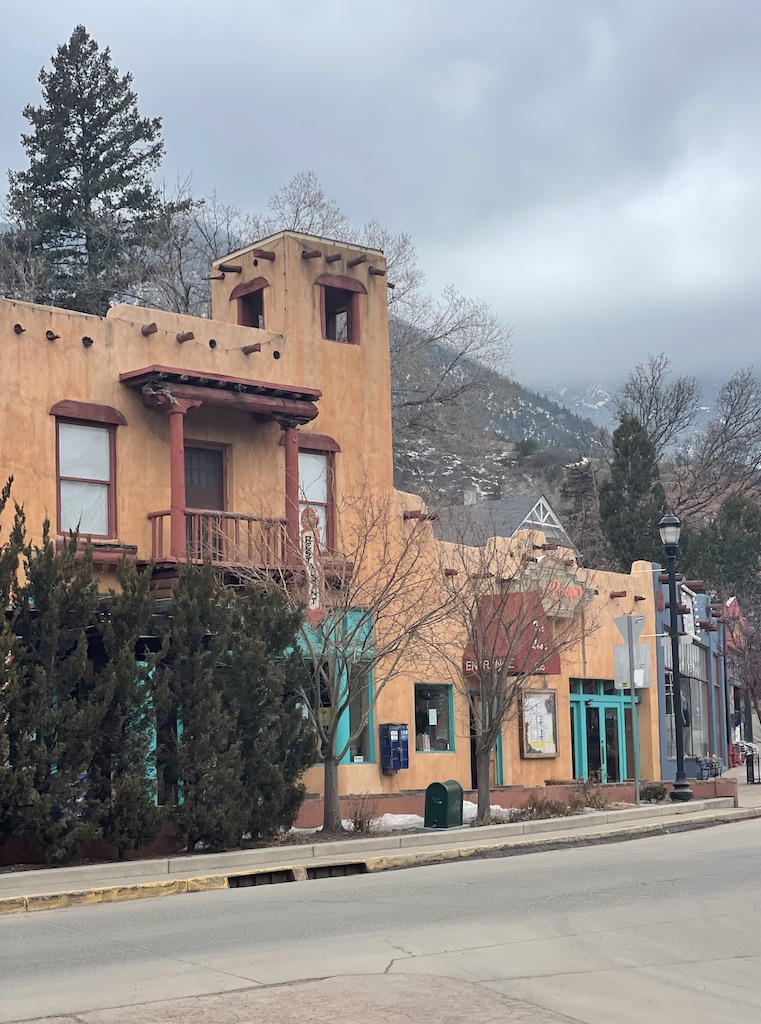 Orange, two story Pueblo building with teal door frames, retail shops along main street with snowy mountains in background on gloomy winter day. Manitou Springs downtown area, Manitou Springs, near Colorado Springs, Colorado.