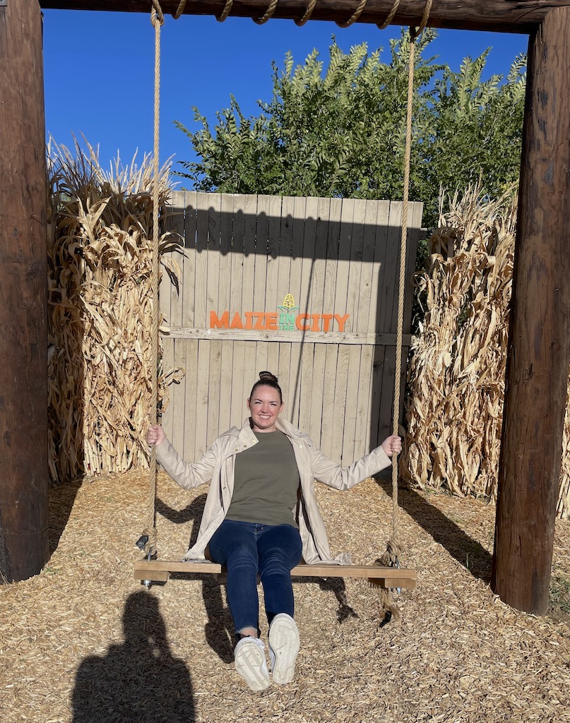 Woman on large, wooden swing smiling, with corn stalks in background on sunny fall day. Pumpkin patch, Maize In The City, Thornton near Denver, Colorado.
