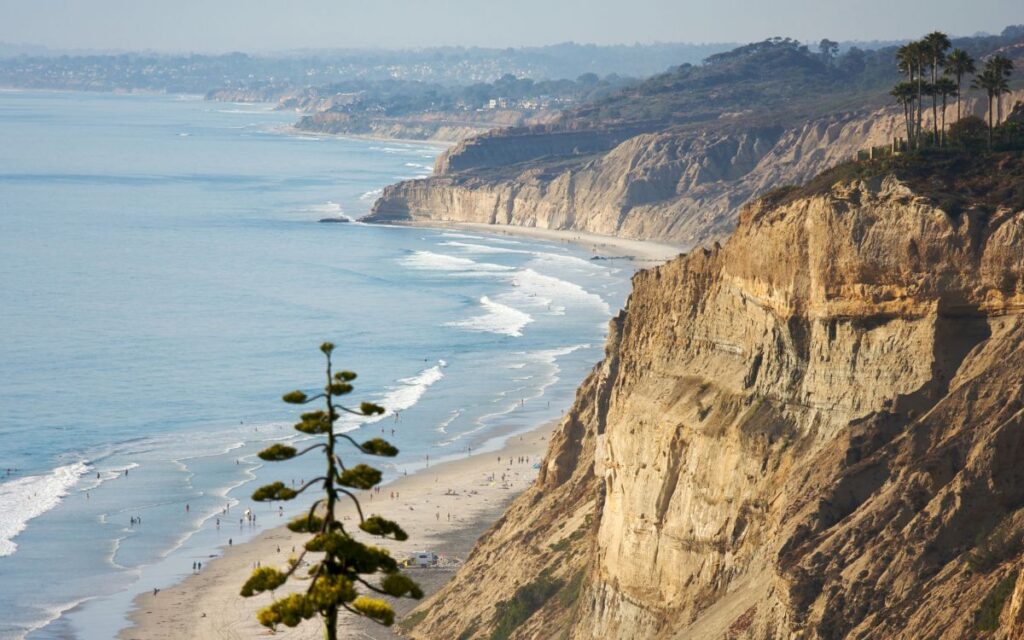 Huge sandy beach cliffs towering over white sand beach dotted with surfers and beach-goers on the Pacific Ocean on hazy, sunny day. Torrey Pines Beach, San Diego, California.