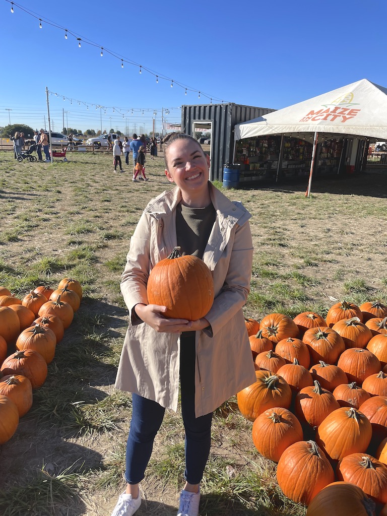 Woman smiling and holding pumpkin in pumpkin patch with string lights and white event tents in background on sunny fall day. Pumpkin patch, Maize In The City, Thornton near Denver, Colorado.