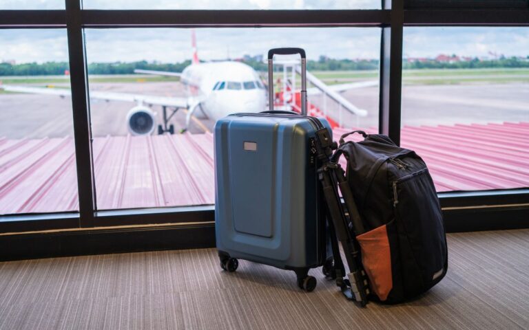 Suitcase and backpack in front of window at airport with large commercial airplane in background on sunny day.