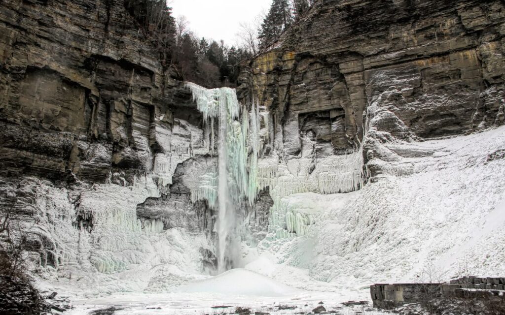 Frozen waterfall "falling" from sheer, stone cliff, surrounded by snow on grey winter day. Taughannock Falls, Taughannock Falls State Park, Ithica, Finger Lakes, Upstate New York.
