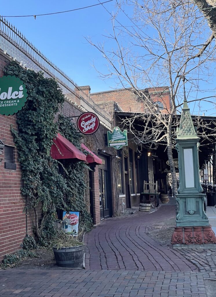 Cobblestone sidewalk next to brick building covered in ivy on sunny day. Shops in Old Market, Tannenbaum Christmas Shop. Tannenbaum Christmas Shop, Old Market, Omaha, Nebraska.