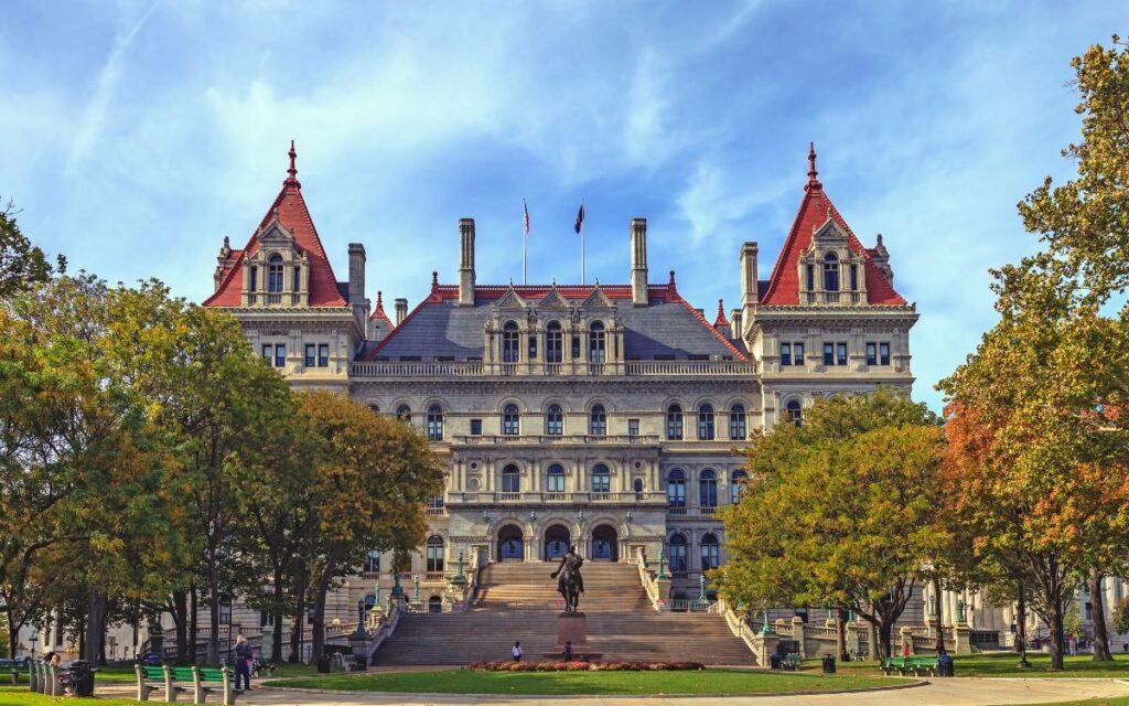 New York's state capitol building in Albany, multi-story building with impressive stonework, orange tiled roof. In front of building is stately statue, mature trees turning in fall, and neat lawn on sunny fall day. New York State Capitol Building, Albany, New York, Upstate.