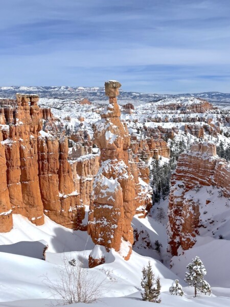 Orange/ red hoodoo canyon covered in snow with snow-covered mountain peaks in background on sunny winter day. Thor's Hammer, Navajo Loop via Queens Loop snowshoeing hike, Bryce Canyon National Park, Utah.