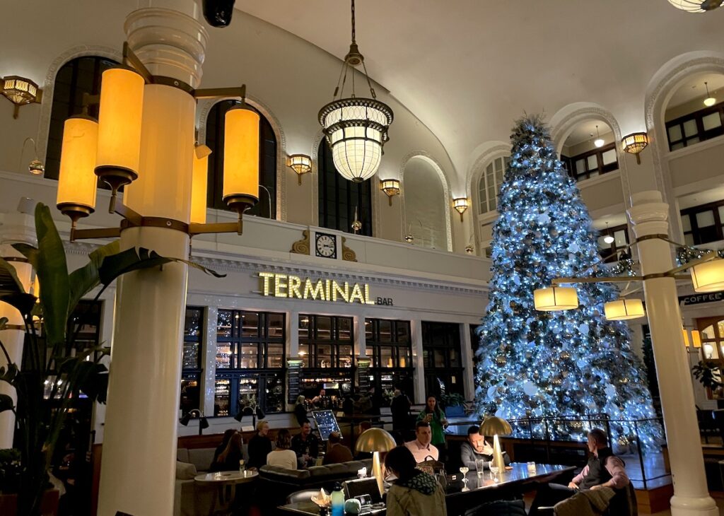 Large Christmas tree with blue lights in chic, art-deco inspired train station bar, with tall ceilings, ornate lights, and lit sign reading "Terminal Bar". Terminal Bar, Denver Union Station, LoDo, Denver, Colorado.