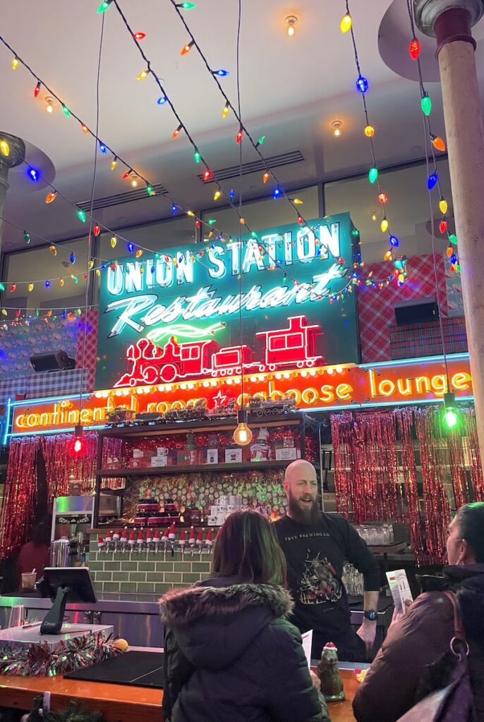 Bar with bartender and patrons under colorful Christmas lights and large neon sign reading "Union Station Restaurant" with train. Miracle at Union Station, Christmas pop-up bar, Union Station, LoDo, Denver, Colorado.
