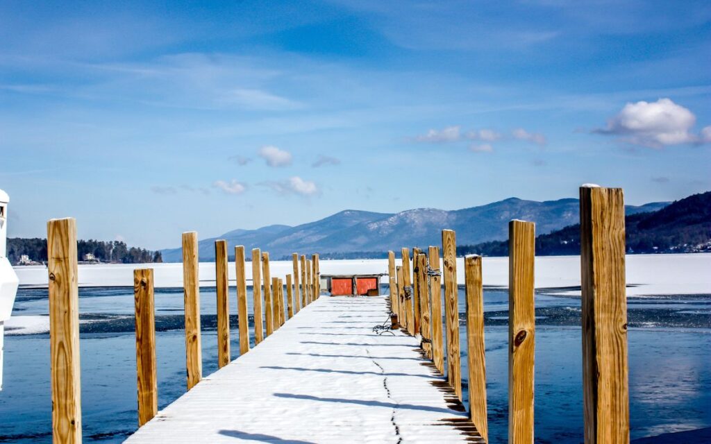Snow-covered wooden dock extending into partially frozen lake with mountain views in background on sunny winter day. Lake George, Adirondack Mountains, Upstate, New York in winter.
