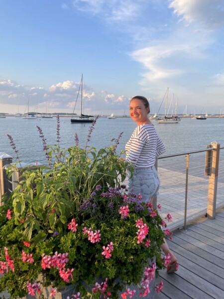 Woman smiling at camera on dock overlooking sailboats and yachts on Sag Harbor during sunny summer evening, with pink and purple flower pots in foreground. Sag Harbor Yacht Yard, Sag Harbor, New York, The Hamptons.