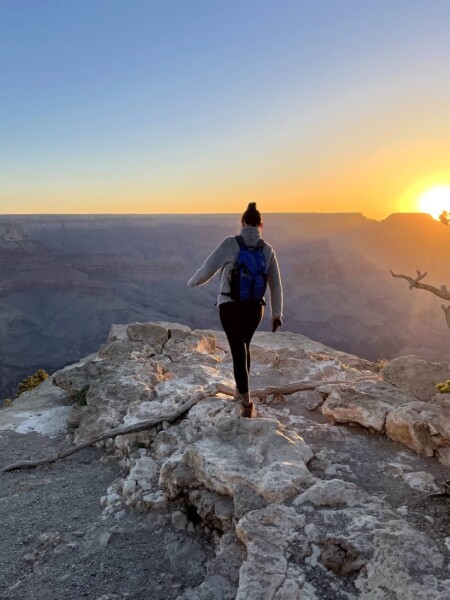 Woman on rocky hiking trail overlooking red, orange, and purple Grand Canyon during sunrise on cloudless, but cold spring morning. Sunrise at Yaki Point, Grand Canyon National Park South Rim, Arizona.