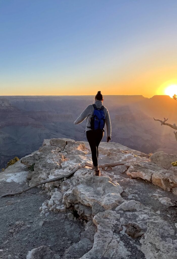 Woman on rocky hiking trail overlooking red, orange, and purple Grand Canyon during sunrise on cloudless, but cold spring morning. Sunrise at Yaki Point, Grand Canyon National Park South Rim, Arizona.