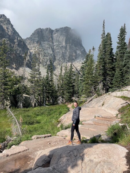 Woman hiking on cloudy summer day next to lush green meadow backed by tall pine trees and huge, rocky mountaintops. Dream and Emerald Lake hikes, Rocky Mountain National Park, Estes Park, Colorado.