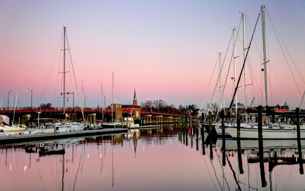Sailboats on calm bay water with large bridge and steepled church on horizon during purple pink sunset. Downtown district, Chesapeake Bay, Annapolis, Maryland.