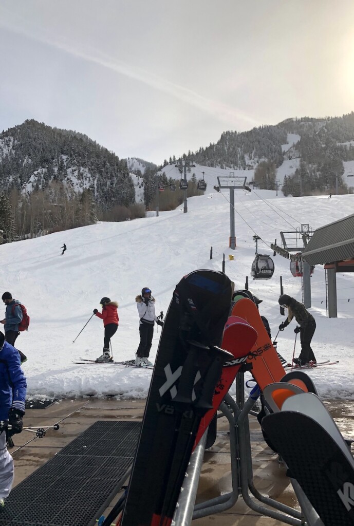 Base of ski resort, ski racks filled with skis and snowboards, gondola heading up snow covered mountain while skiers come down during sunny afternoon in winter. Aspen Mountain (Ajax), Aspen in winter, Colorado.