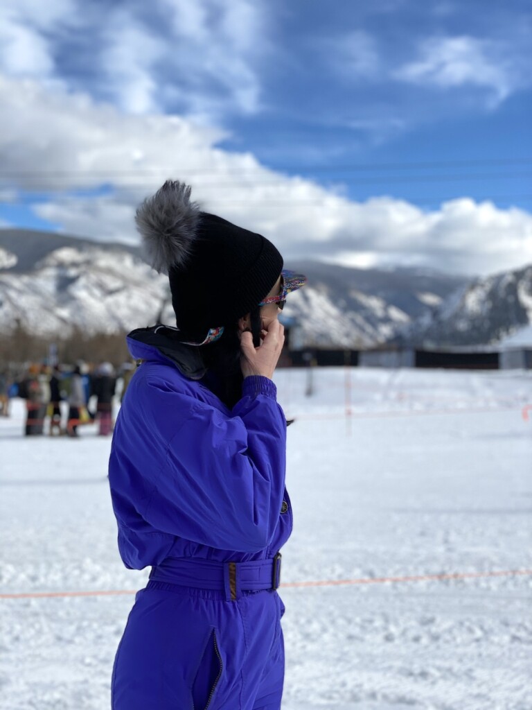 Woman in purple snowsuit and black hat standing in snow on skill hill with snow-covered mountains in background on sunny winter day. Aspen Snowmass Ski Resort, Aspen X-Games, Snowmass Village, Colorado.