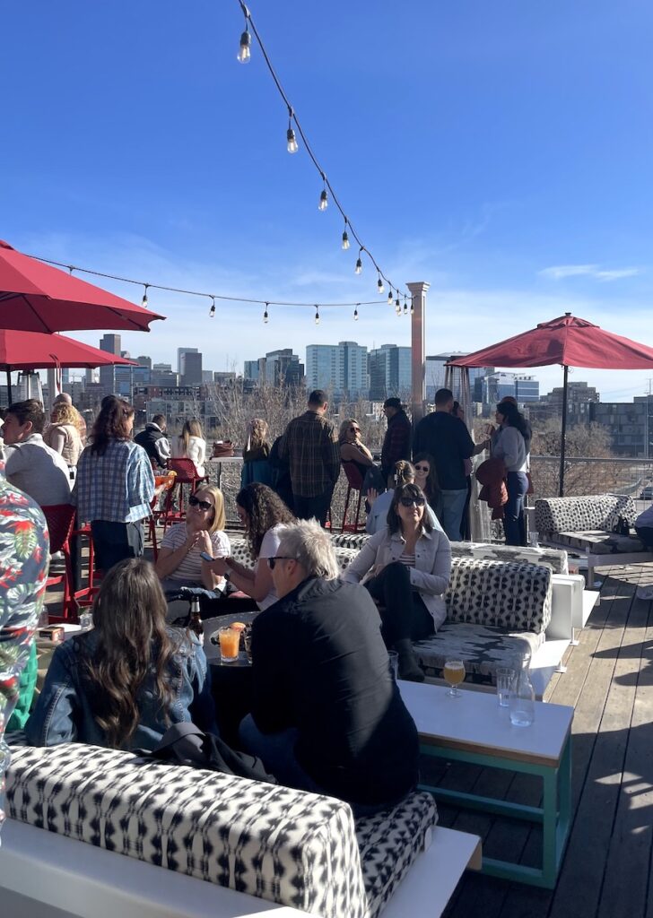 Outdoor rooftop deck full people having drinks and hanging out under string lights on sunny, warm winter day with Denver skyline views in background. Avanti Food & Beverage, The Highlands, Denver, Colorado.