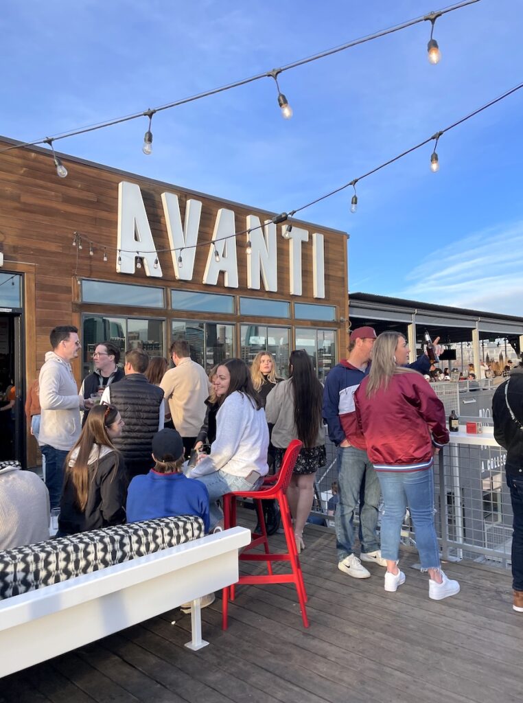 Outdoor rooftop deck full people having drinks and hanging out under string lights and large building sign reading "AVANTI" on sunny, warm winter day. Avanti Food & Beverage, The Highlands, Denver, Colorado.