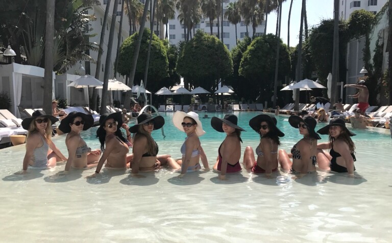 Group of women sitting in shallow end of pool surrounding bride-to-be during bachelorette party, in background are pool cabanas and palm trees. Pool at Delano South Beach, South Beach, Miami, Florida.