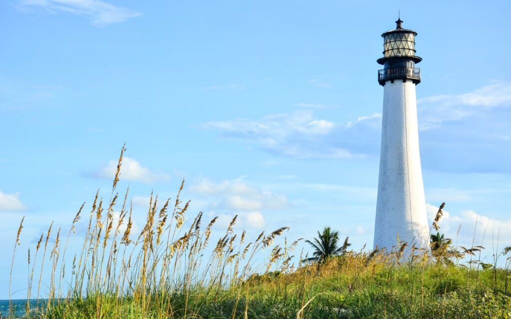 White lighthouse on edge of water with greenery in foreground on sunny day. Cape Florida Lighthouse, Bill Baggs Cape Florida State Park, Key Biscayne, Miami, Florida.