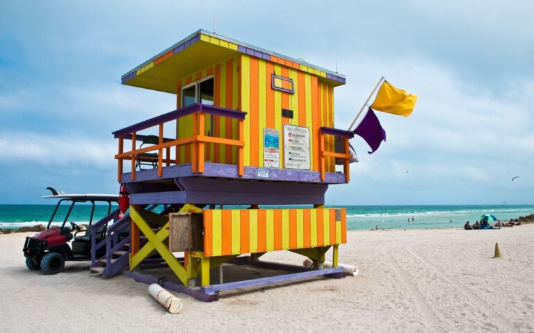 Yellow, orange, and purple striped lifeguard stand on beach with bright blue water and beach-goers in background. Miami Beach, South Beach, Miami, Florida.