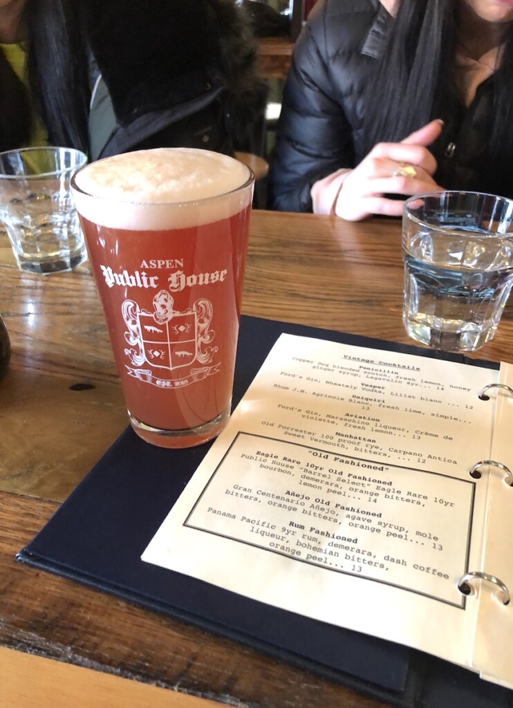 Pink-hued craft beer in clear glass reading "Aspen Public House" next to open menu at restaurant table. Aspen Public House, Aspen, Colorado.