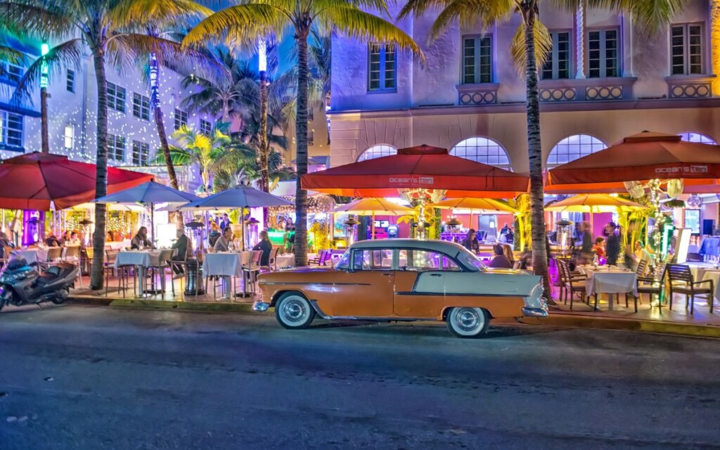 Old, 1920's car in front of restaurant with sidewalk seating under palm trees and buildings at night. Ocean's Ten, Ocean Drive, South Beach, Miami, Florida.