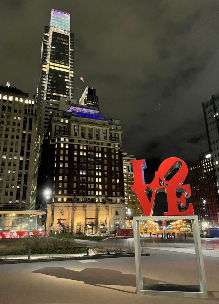 Nighttime view of city skyline lit up with small red statue in foreground reading "Love". Love Statue, Downtown Philadelphia, Pennsylvania.