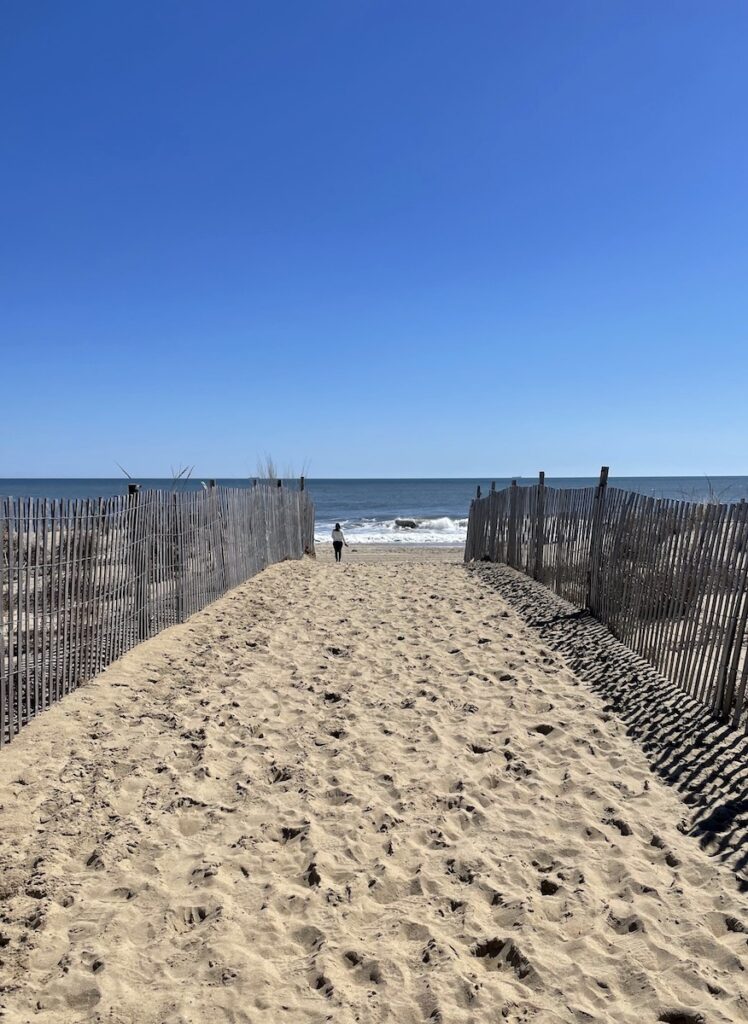 Path of golden sand leading to sandy beach on Atlantic Ocean with person in distance on cloudless day. Rehoboth Beach, Delaware.