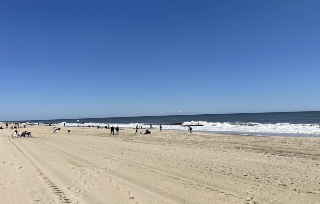 Silhouette of beach-goers on golden sand beach next to waves of Atlantic Ocean on cloud-less summer day. Rehoboth Beach, Rehoboth Beach, Delaware.