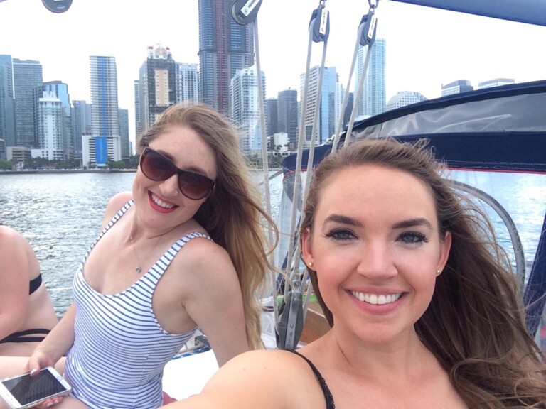 Two women smiling at camera on sailboat with Biscayne Bay and Downtown Miami skyline in background during sailboat charter. Biscayne Bay, Downtown Miami, Miami, Florida.