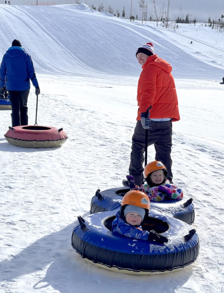 Man in orange jacket pulling young kids in tandem tubs in snow with tubing snow course in background.