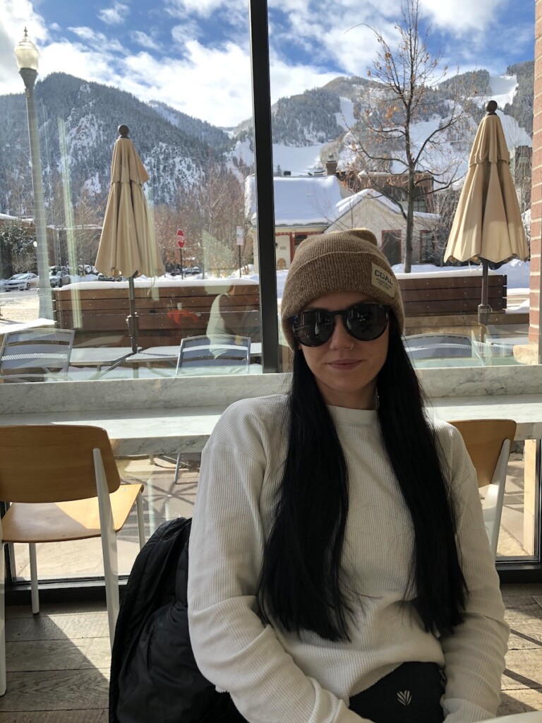 Woman sitting in cafe with large glass windows in background overlooking snow-covered ski resort on sunny winter day. Spring Cafe, downtown Aspen, Colorado.
