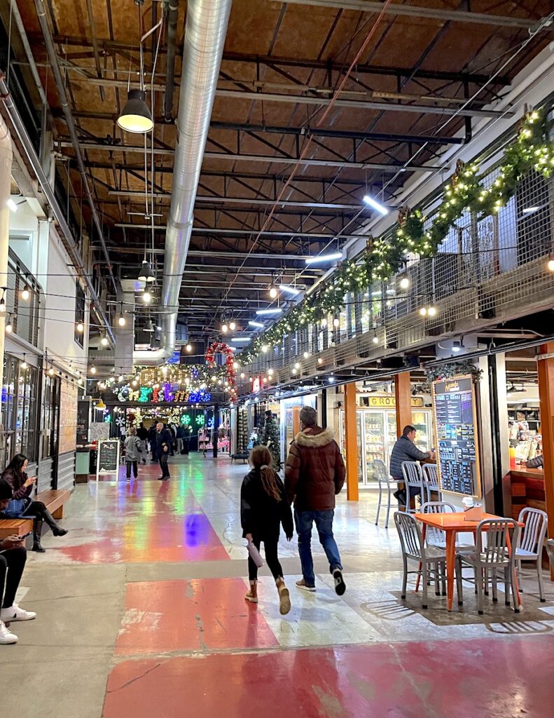 People walking through two story food hall with huge, open industrial ceilings, food vendors are lit up and surrounded by Christmas lights. The Stanley Marketplace, Aurora, near Central Park, Denver, Colorado.