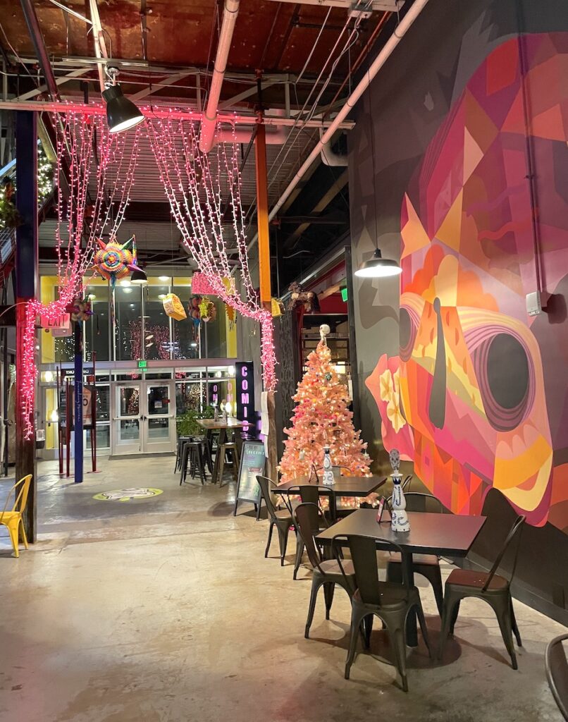 Pink, yellow, and orange colorful wall mural on interior wall in food market next to cafe tables, and pink decor. Comida, The Stanley Marketplace, Aurora, near Central Park, Denver, Colorado.