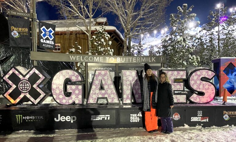 Two women in front of huge sign reading "X GAMES" with snow-covered pine trees and mountains in background. X Games, Buttermilk Ski Resort, Aspen, Colorado.