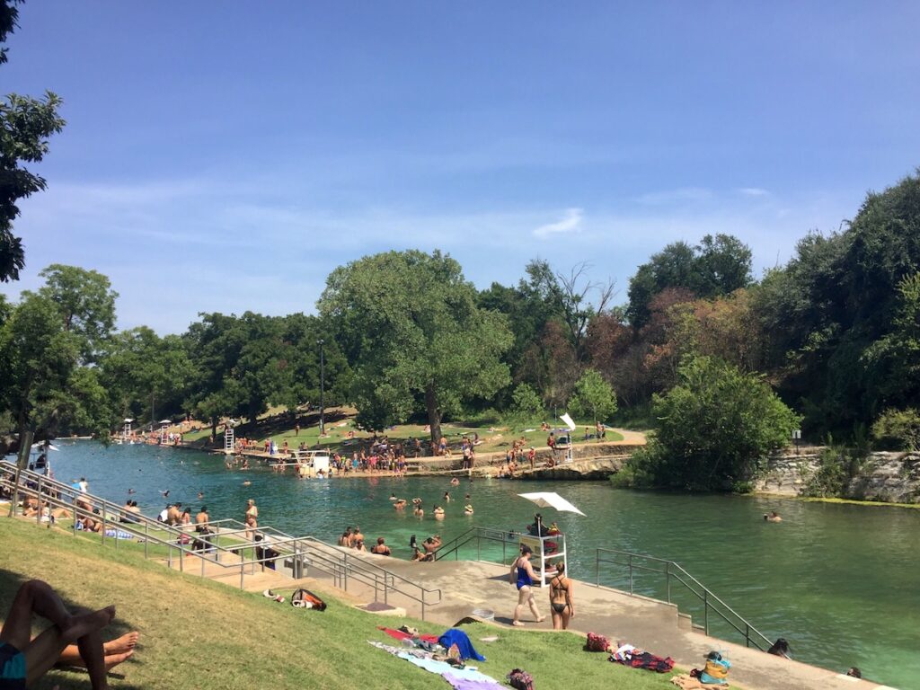 People in outdoor natural spring pool surrounded by green lawn and green trees on sunny summer day. Barton Springs Pool, Austin, Texas.