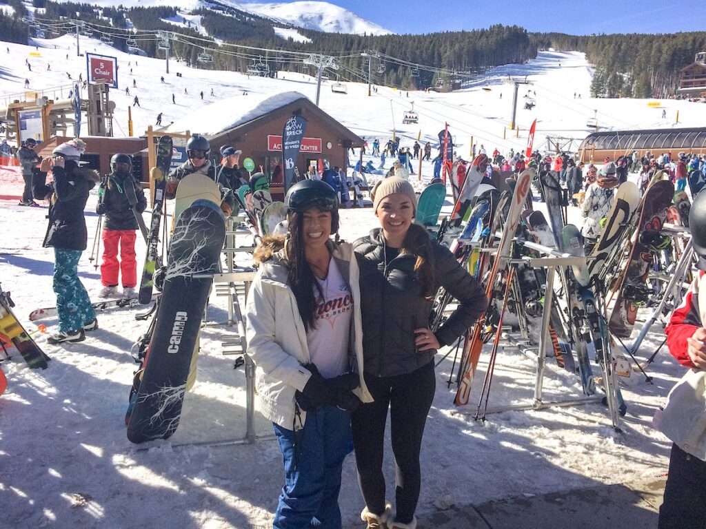 Two women smiling in front of ski racks and ski lifts at base of mountain ski resort. TBar, base of Peak 8, Breckenridge Ski Resort, Breckenridge, Colorado.