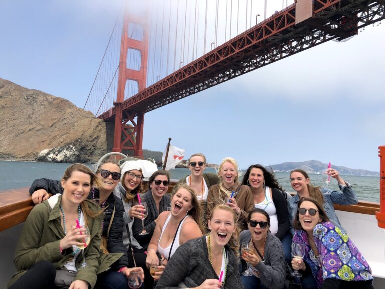 Group of woman on boat under Golden Gate Bridge smiling at camera and holding wine glasses. Wine boat tour, bachelorette party, San Francisco, California.