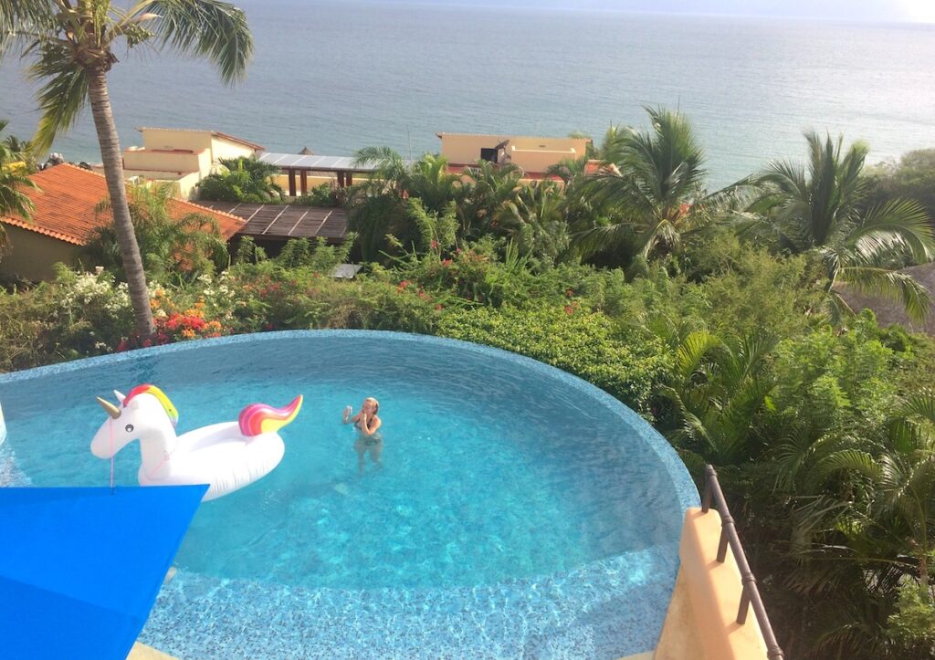 Woman and giant unicorn floatie in infinity pool overlooking lush greenery, palm trees, and pacific ocean on sunny winter day. Vacation home near Puerto Vallarta, Mexico.