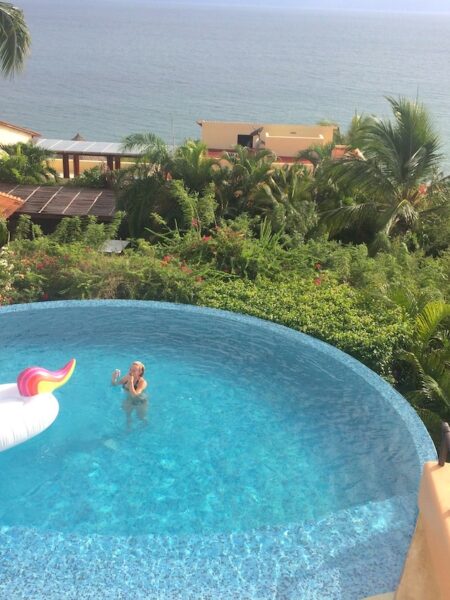 Woman and giant unicorn floatie in infinity pool overlooking lush greenery, palm trees, and pacific ocean on sunny winter day. Vacation home near Puerto Vallarta, Mexico.
