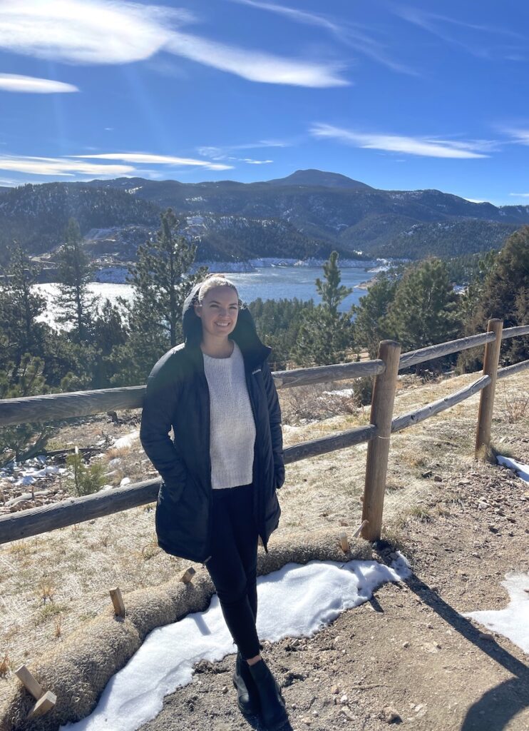 Woman leaning against wooden fence, overlooking reservoir lake under mountains on sunny winter day. Gross Reservoir, Boulder County, Colorado.