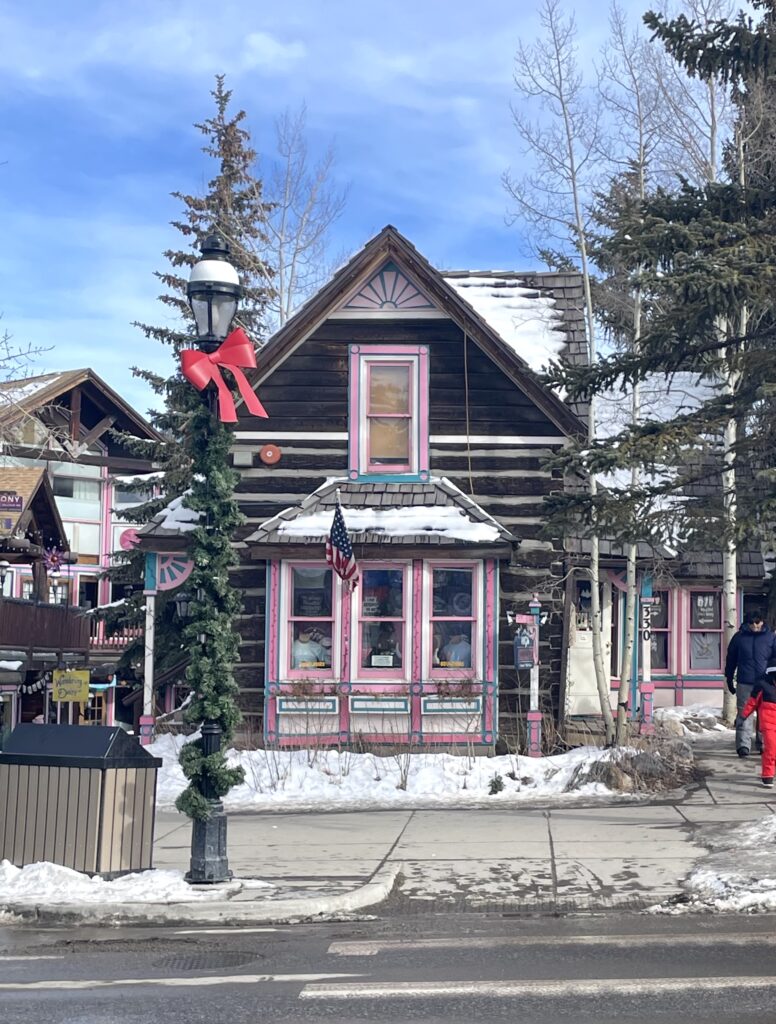 Small wooden house with pink and blue trim surrounded by snow and pine trees on Main Street on sunny winter day. Breckenridge Historic District, Main Street, Breckenridge, Colorado.