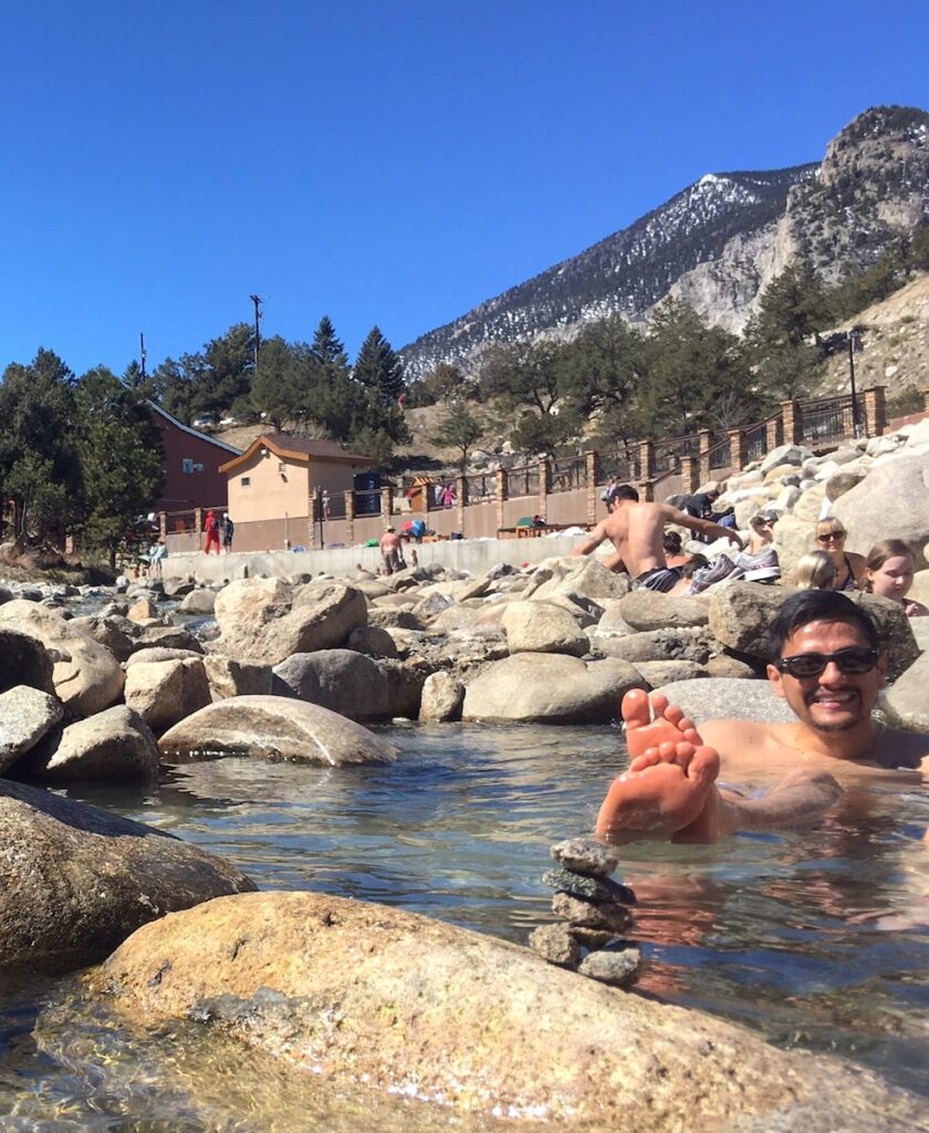 Man soaking in creekside hot springs with mountain and resort buildings in background on sunny spring day. Mt. Princeton Hot Springs Resort, Buena Vista, Colorado.