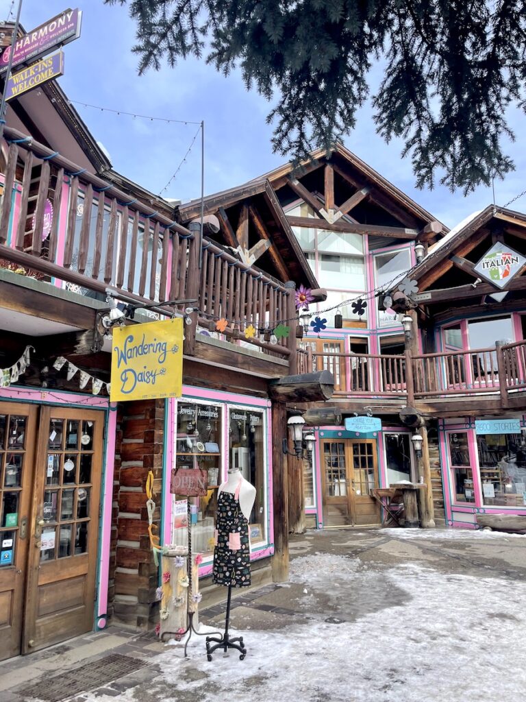 Businesses in wooden buildings with pink and blue trim surrounded by pine trees and snow on sunny winter day. Breckenridge Historic District, Main Street, Breckenridge, Colorado.