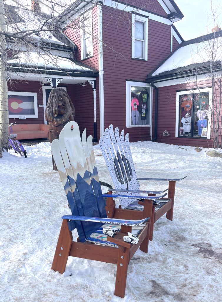 Two Adirondack chairs made of old skis on snowy front lawn in front of red building on sunny day in winter. Breckenridge Historic District, Main Street, Breckenridge, Colorado.